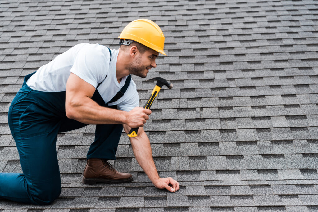 handsome repairman in helmet holding hammer while repairing roof in house